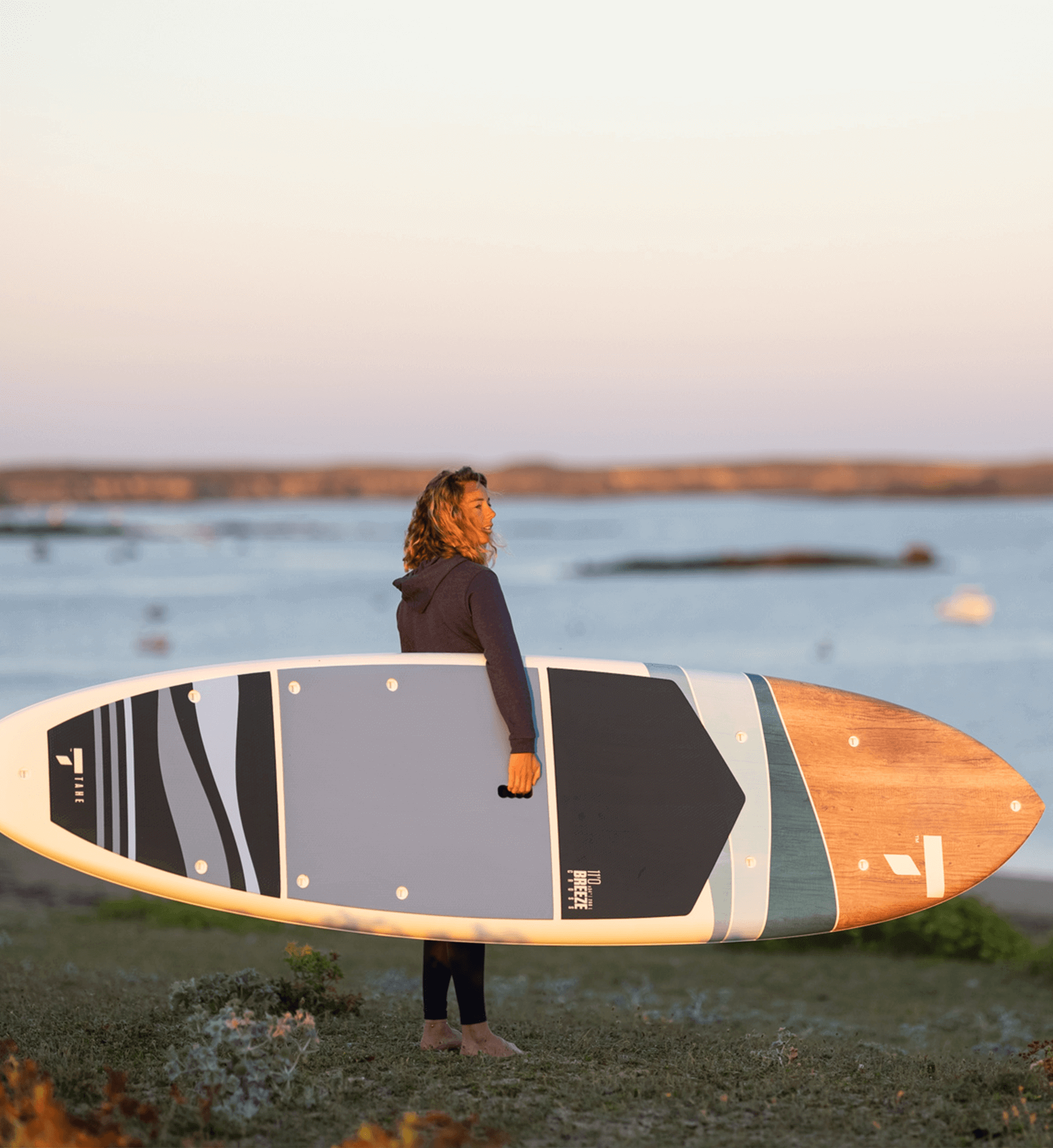 Stylish paddler in a blue hoodie carrying a TAHE SUP with a classic blue and wood design, overlooking the ocean from a grassy dune.