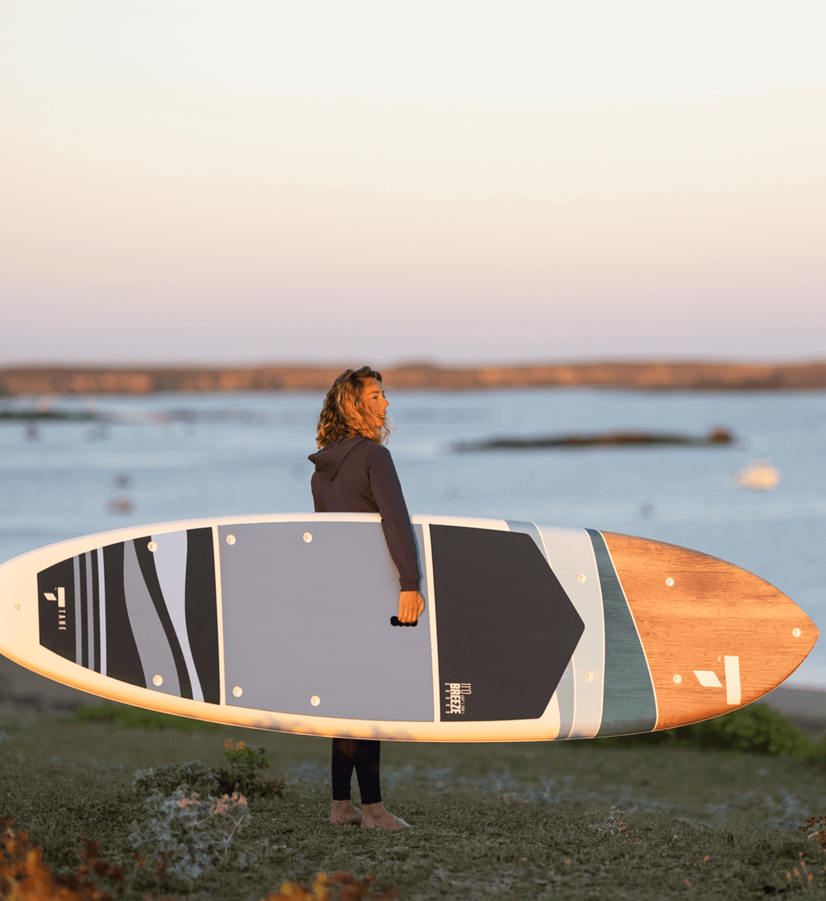 Stylish paddler in a blue hoodie carrying a TAHE SUP with a classic blue and wood design, overlooking the ocean from a grassy dune.