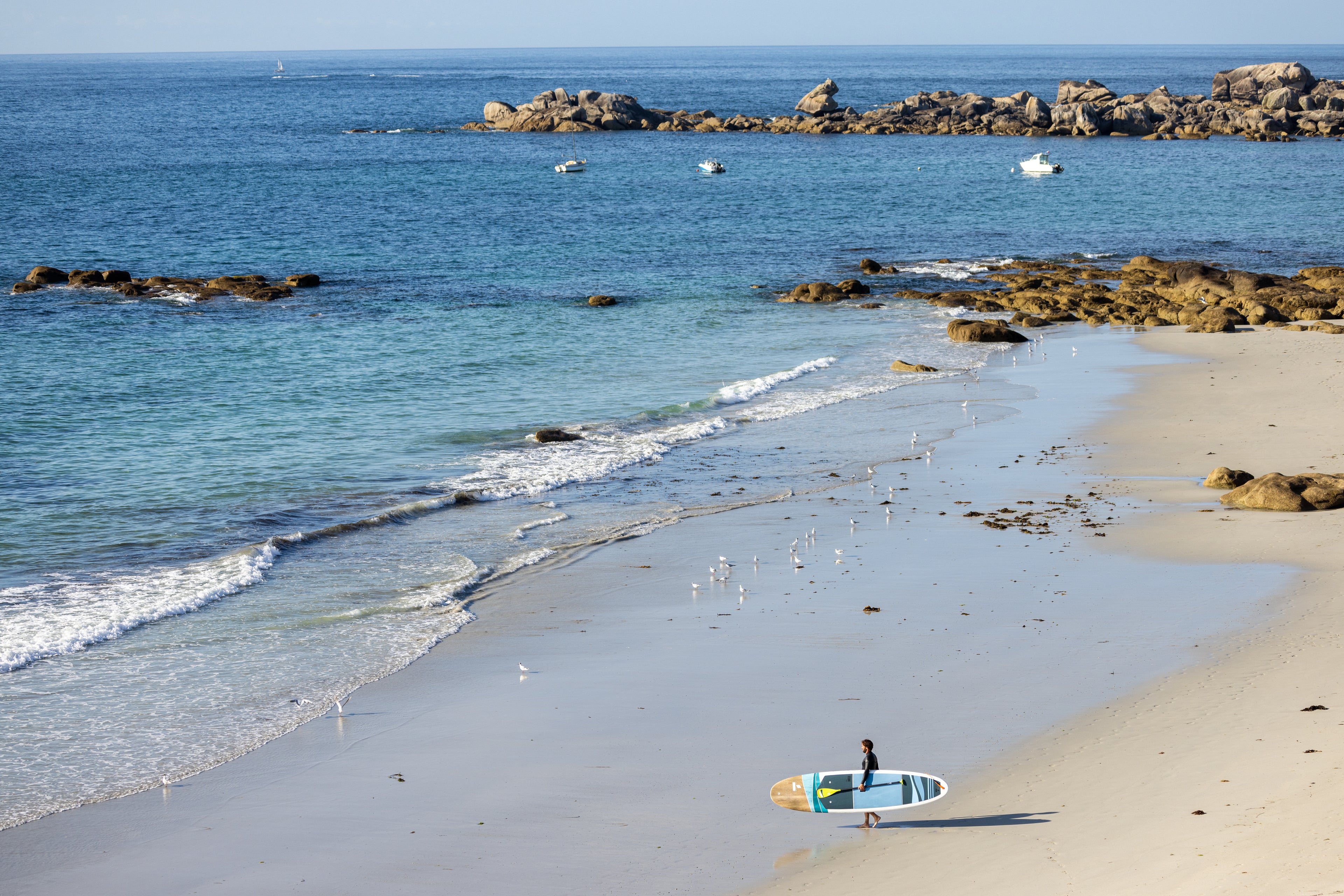 Man carrying a TAHE paddleboard under his arm on a beach facing the ocean.